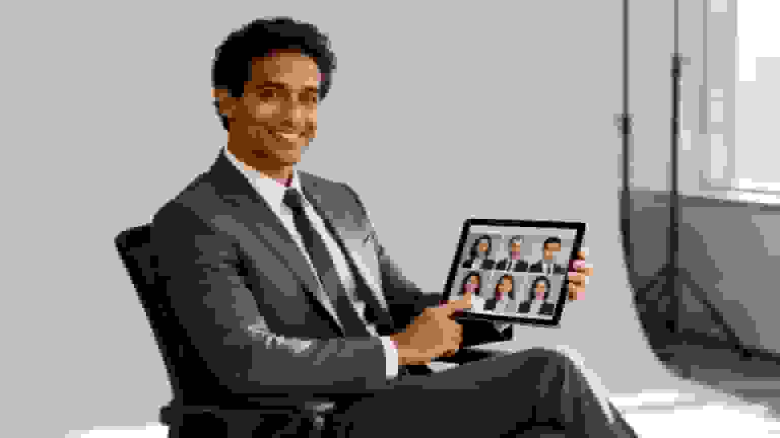 Smiling businessman in a suit holds a tablet displaying a grid of diverse professional headshots.