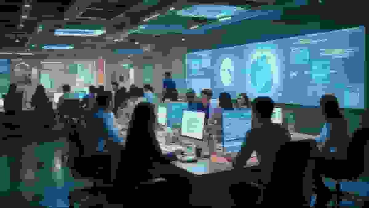 An open-plan office with people working at desks in front of a large data screen