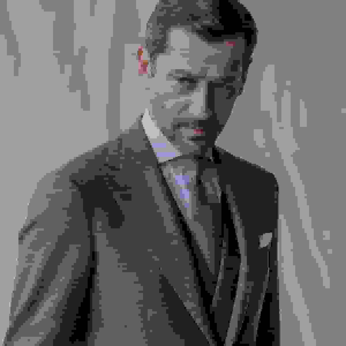 A man in a grey wool suit, light blue shirt and brown tie against a soft neutral backdrop