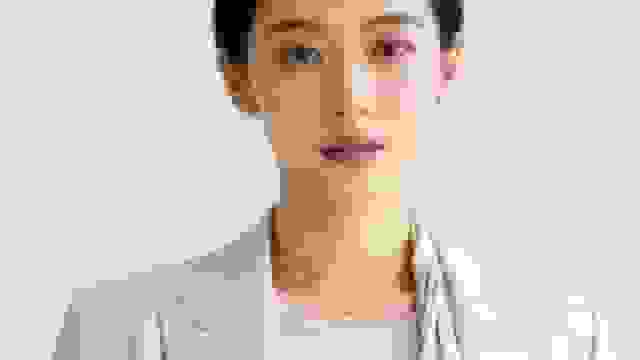 Close-up of an Asian woman in a light blazer, wearing pearl earrings and a necklace.