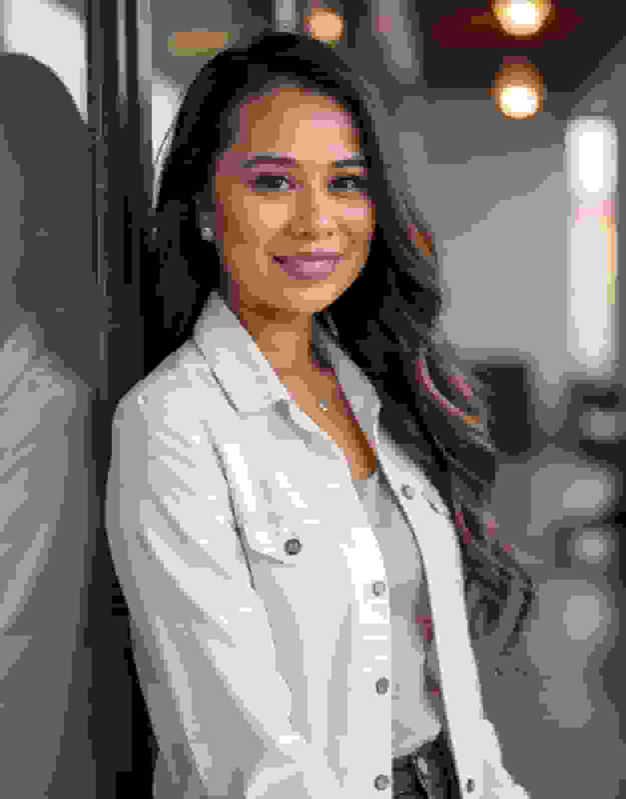 Casual headshot of a smiling woman with long wavy hair in a white denim jacket over a pink top, leaning against a glass wall in an office
