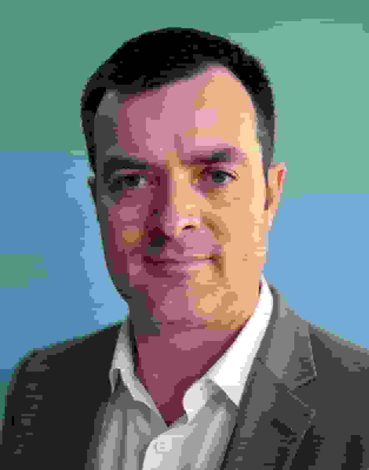 Business casual headshot of a man in a grey blazer and an open-collar white shirt on a plain blue studio backdrop
