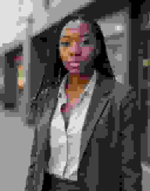 Headshot of a woman with long braids worn down over her shoulders, in a charcoal blazer, on a blurred city street